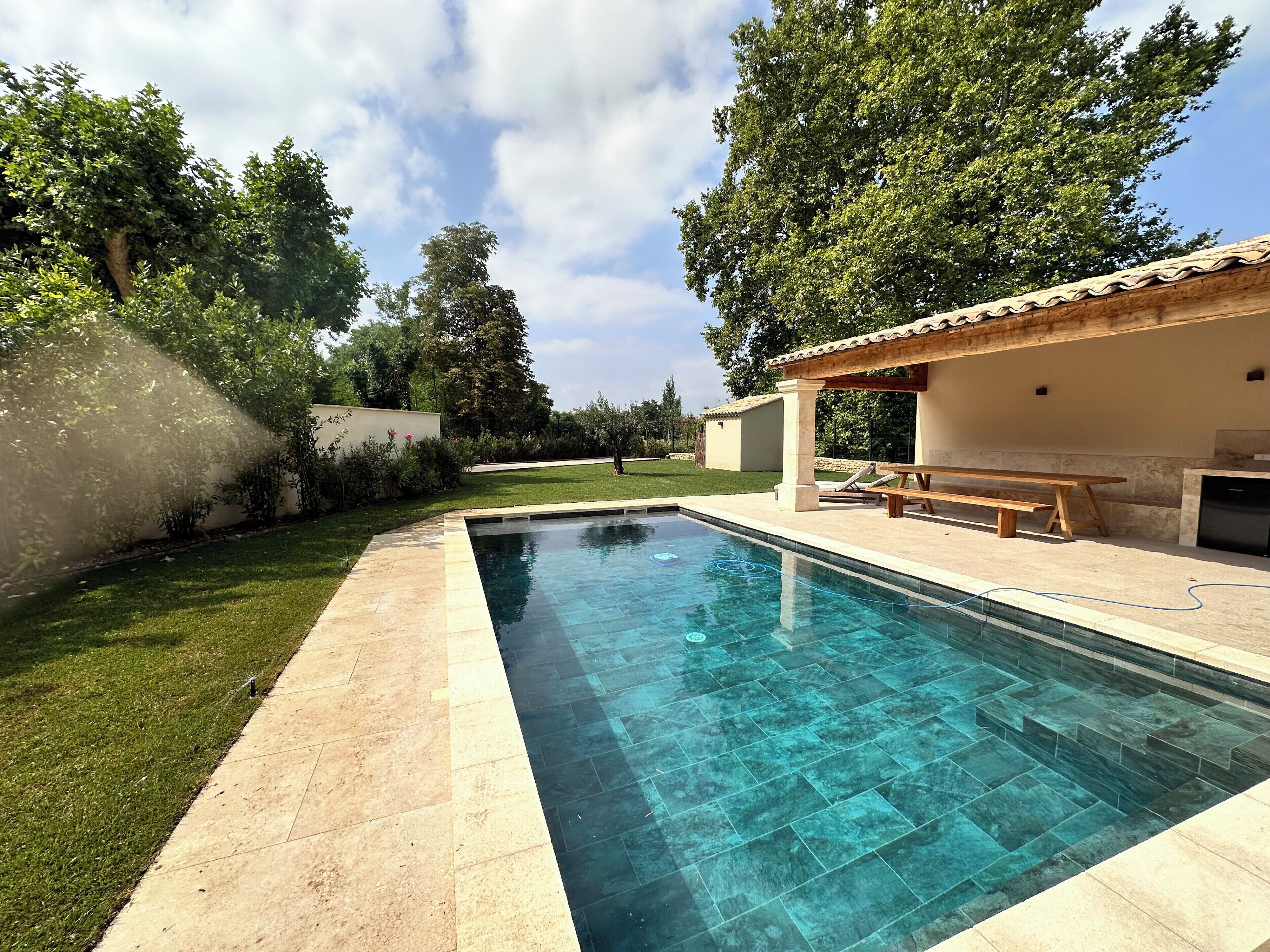 A swimming pool with a tiled deck, a pergola with seating, and a grassy area with trees.