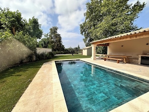 A swimming pool with a tiled deck, a pergola with seating, and a grassy area with trees.