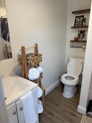 Un baño con un lavamanos blanco, un estante de madera con cestas, un inodoro y un espejo.