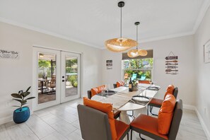 Un comedor con una mesa dispuesta para cuatro personas, sillas con cojines naranja, una lámpara colgante de fibras naturales y vistas a un patio exterior a través de puertas de vidrio.