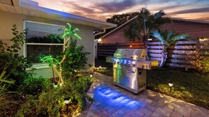 Eine Terrasse mit Grill aus Edelstahl, Holzzaun und ein Haus mit Fenster.