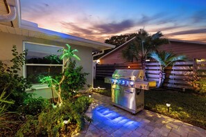 Eine Terrasse mit Grill aus Edelstahl, Holzzaun und ein Haus mit Fenster.