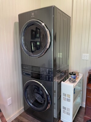 A stacked washer and dryer unit with a control panel, a small shelf with a container, and a power outlet on the wall. - Equestrian cottage— country living in the city (Aiken)