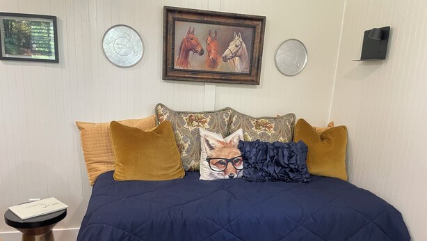 A bed with a dark blue quilted bedspread, multiple pillows in various colors and patterns, a framed painting of horses, and a small table with a book beside the bed. - Equestrian cottage— country living in the city (Aiken)