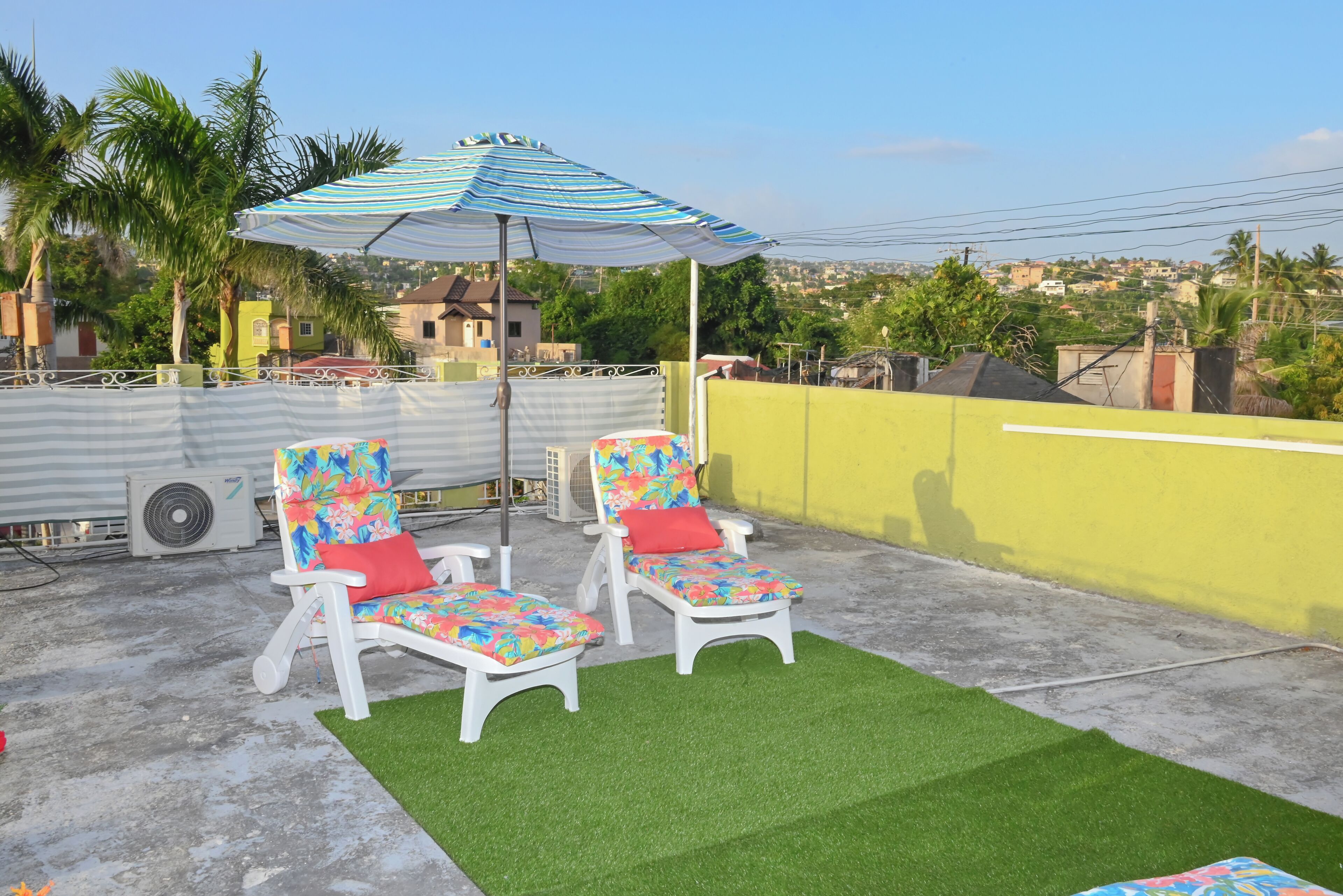 A rooftop terrace with two white lounge chairs, a striped umbrella, and a view of a residential area.