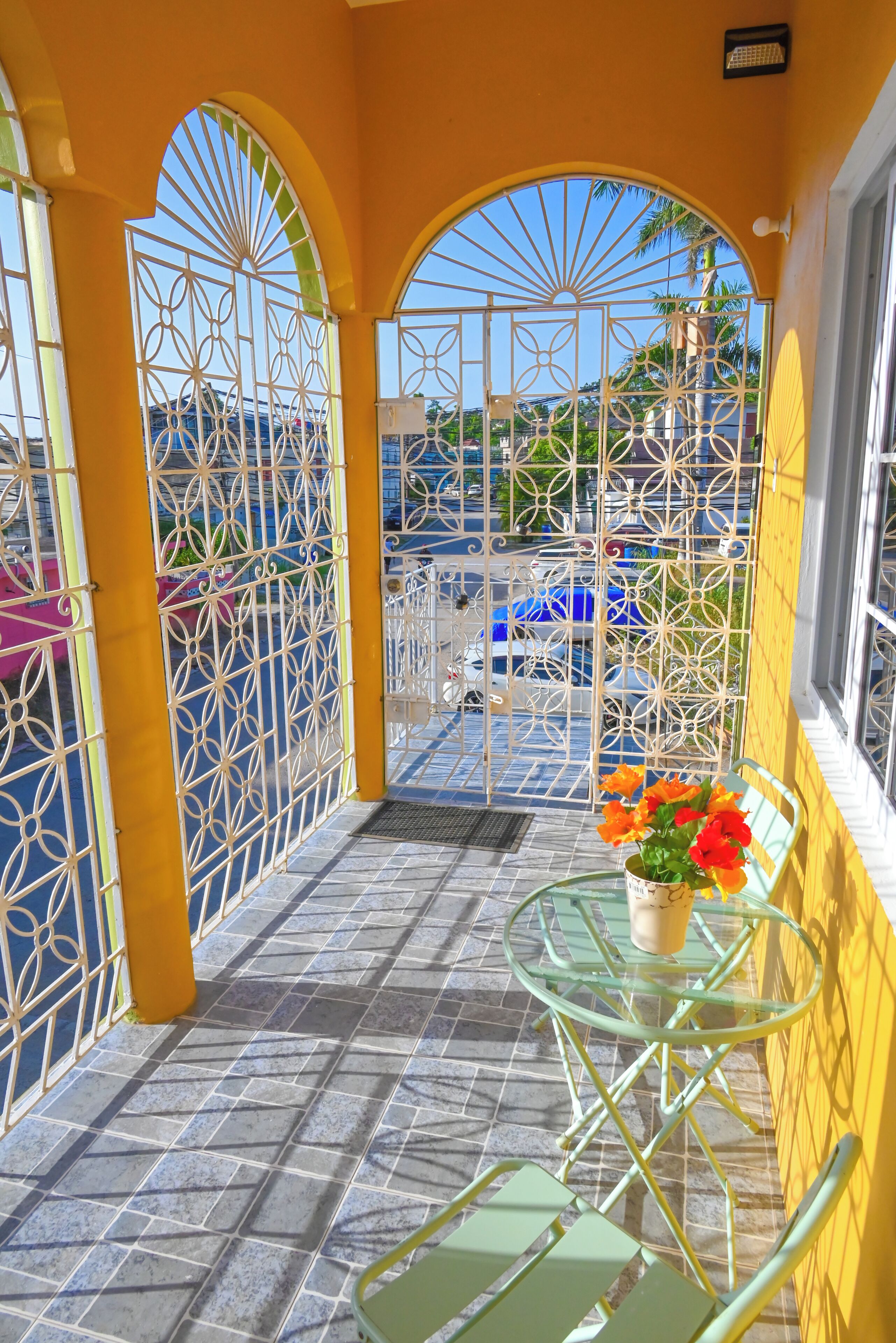 Un balcon aux murs jaunes, une table en verre, une chaise et un pot de fleurs.