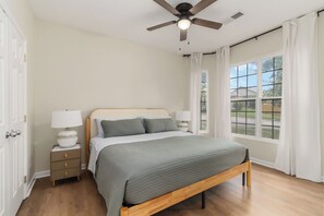 Un dormitorio con cama, mesitas de noche, ventilador de techo y ventana con cortinas.