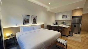 Ein Hotelzimmer mit Bett, Nachttisch, Lampe und einem kleinen Schreibtisch mit Stuhl. Es gibt eine Küchenzeile mit Kühlschrank, Herd und Barhocker.