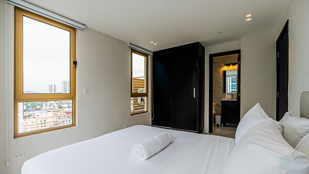 Quarto de hotel com uma cama grande, banheiro com espelho e vista para a paisagem urbana pela janela.