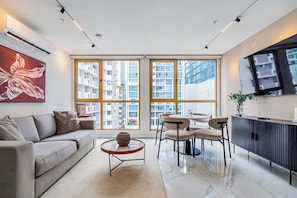 Un salon moderne comprenant un canapé, une table basse et des fauteuils, de grandes fenêtres offrant une vue sur la ville, un téléviseur à écran plat et un meuble de rangement.
