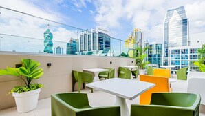 Eine Dachterrasse mit grünen und weißen Sitzgelegenheiten, Pflanzgefäßen und Blick auf eine Stadtlandschaft.