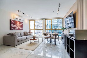 Una sala moderna con sofá, una mesa de centro y un comedor con sillas. Hay un ventanal con vistas a edificios.