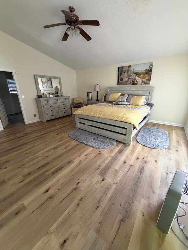 A bedroom with a wooden floor, a bed with a headboard, a ceiling fan, a dresser, a chair, and a painting on the wall.