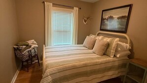 Un dormitorio con una cama, una ventana con cortinas, una mesita, una silla, un cuadro en la pared y una lámpara de techo.