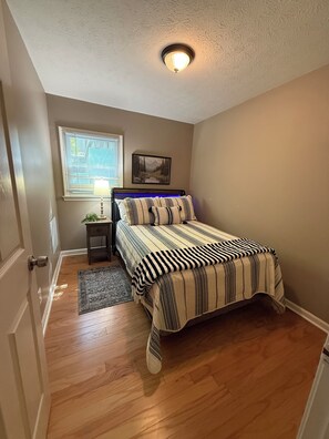 A bedroom with a bed, a nightstand, a lamp, a window with blinds, a painting on the wall, and a ceiling light fixture.