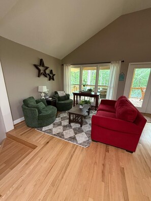 A spacious living room with a wooden floor, green and red sofas, a coffee table, and a large window offering a view of a deck.