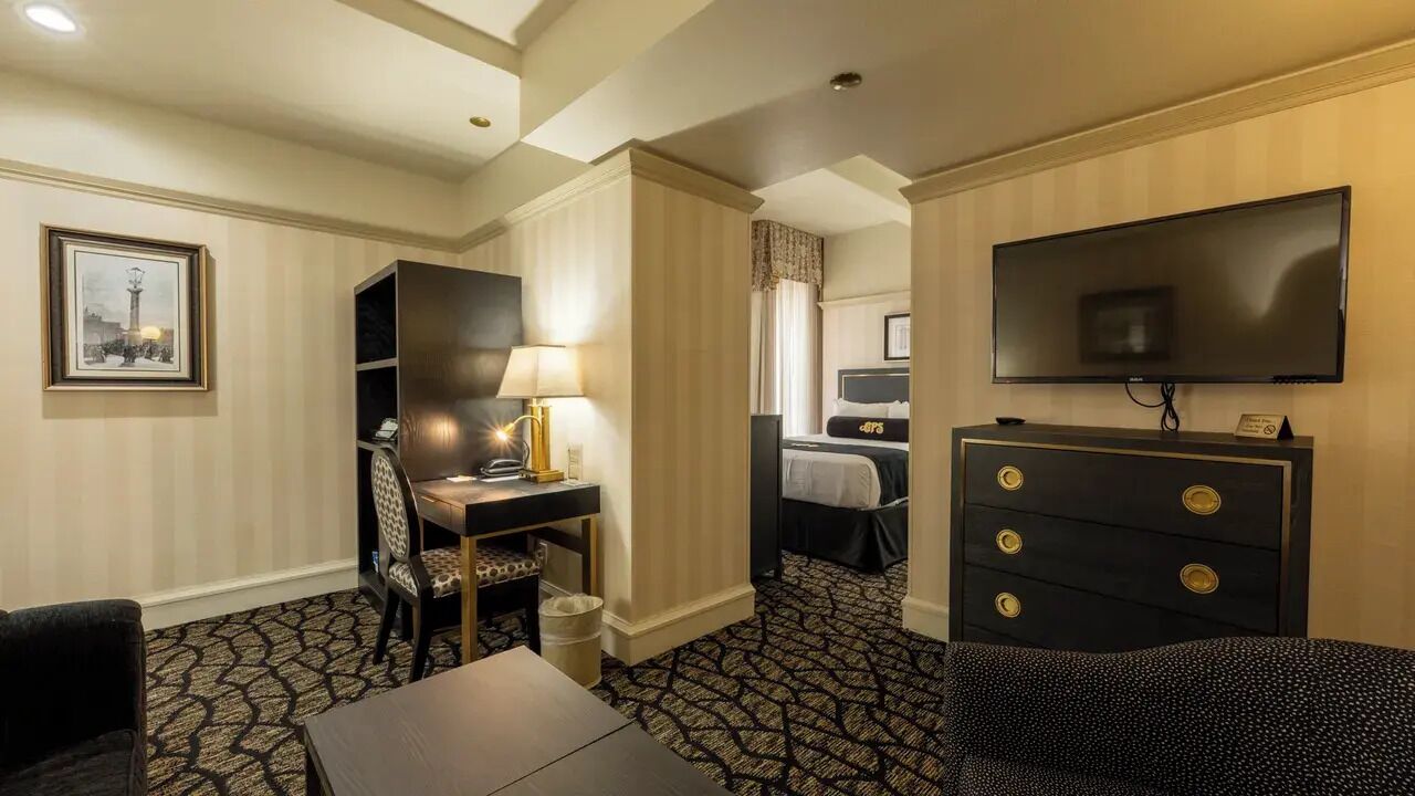 A hotel room with a desk, chair, television, dresser, and a patterned carpet.