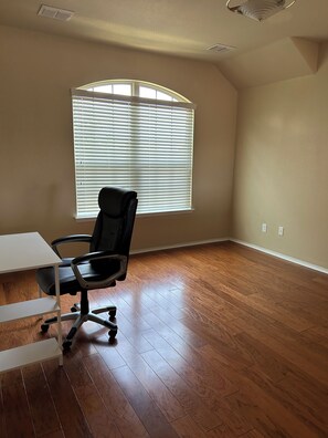 A room with a wooden floor, a black office chair, a white desk, and a window with blinds.