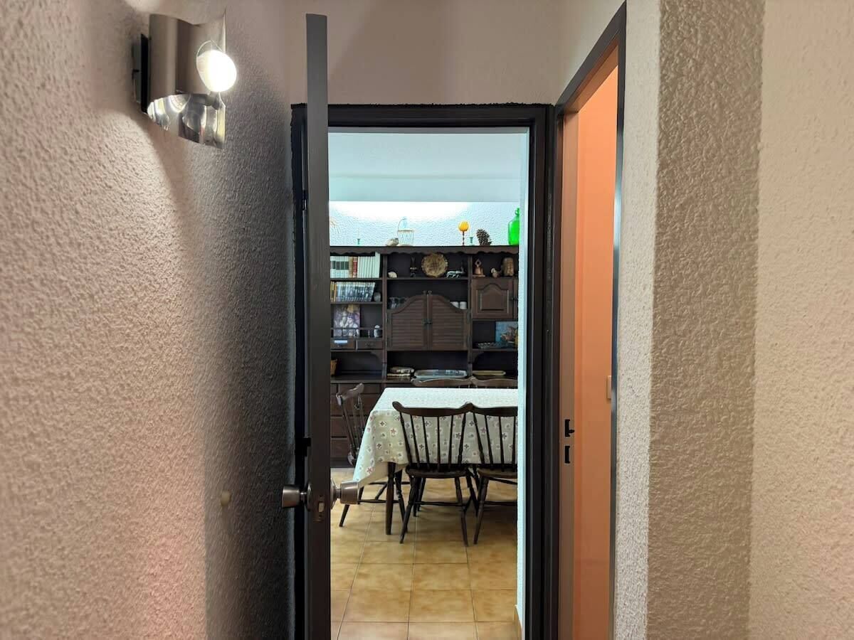 An open door leading to a dining area with a table and chairs, a bookshelf, and a kitchen area with cabinets and a countertop.