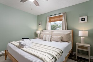 A neatly made bed with a striped blanket, two bedside lamps, a window with curtains, and a small bedside table with a lamp. - Pet-Friendly Oak Island Home W/ Fenced Yard &Fire Pit (Oak Island)