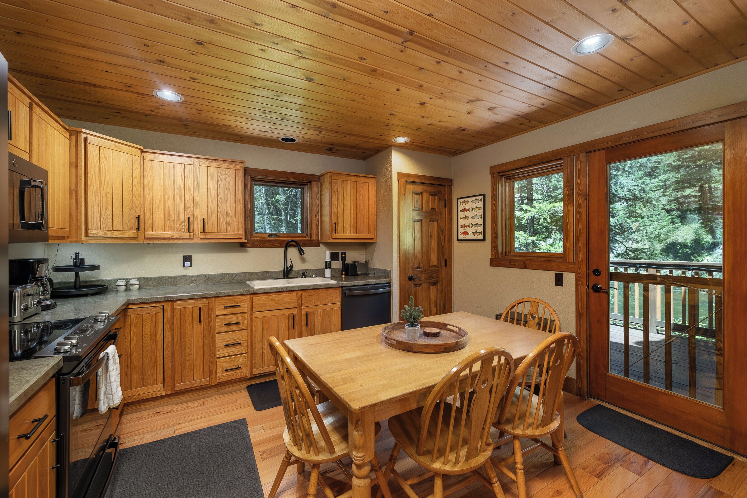 A wooden kitchen with a dining table and chairs, wooden cabinets, and a view of the outdoors through large windows.