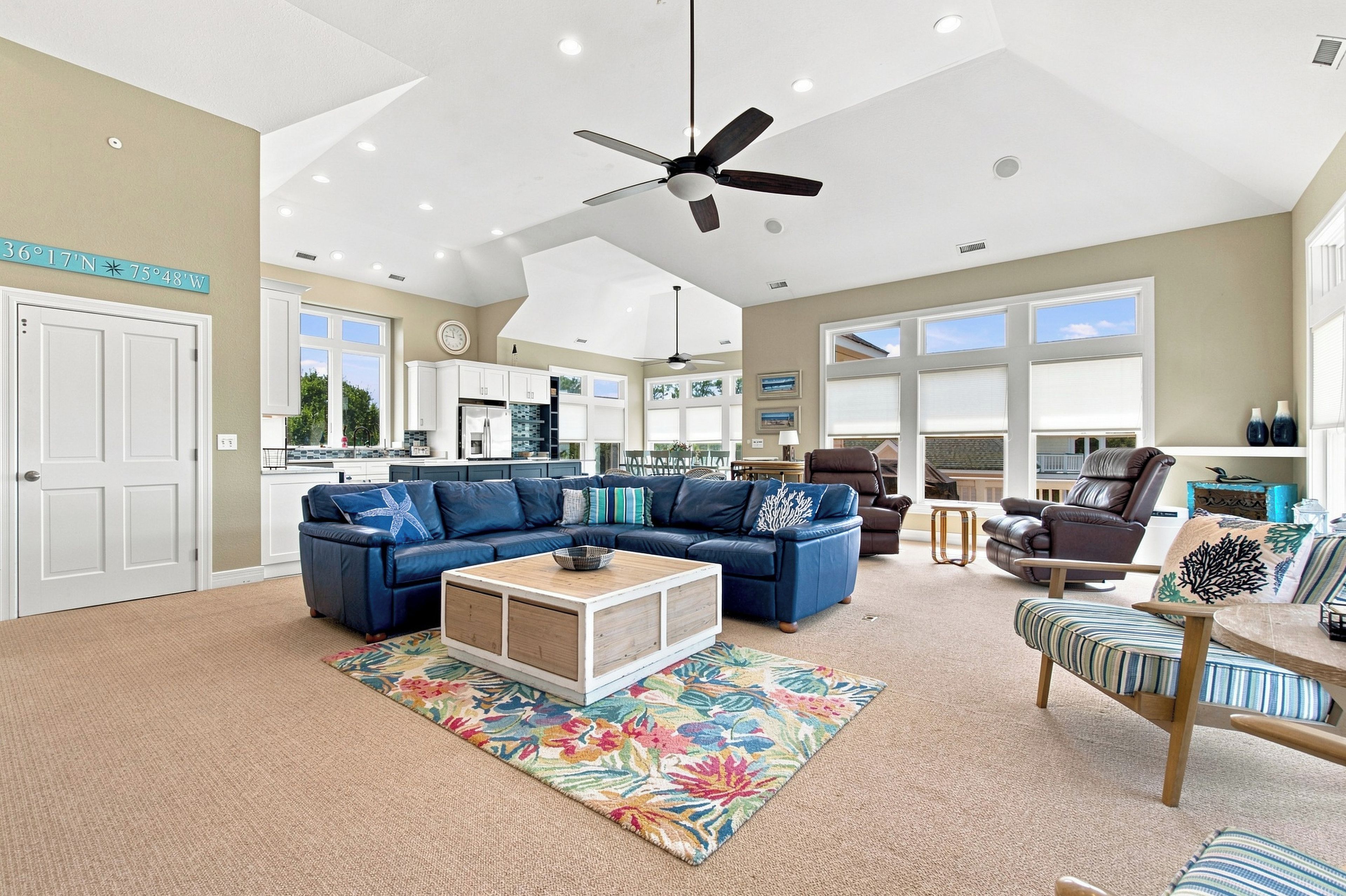 A spacious living room with a ceiling fan, a large sofa set, a coffee table, and a view of the kitchen and dining area.