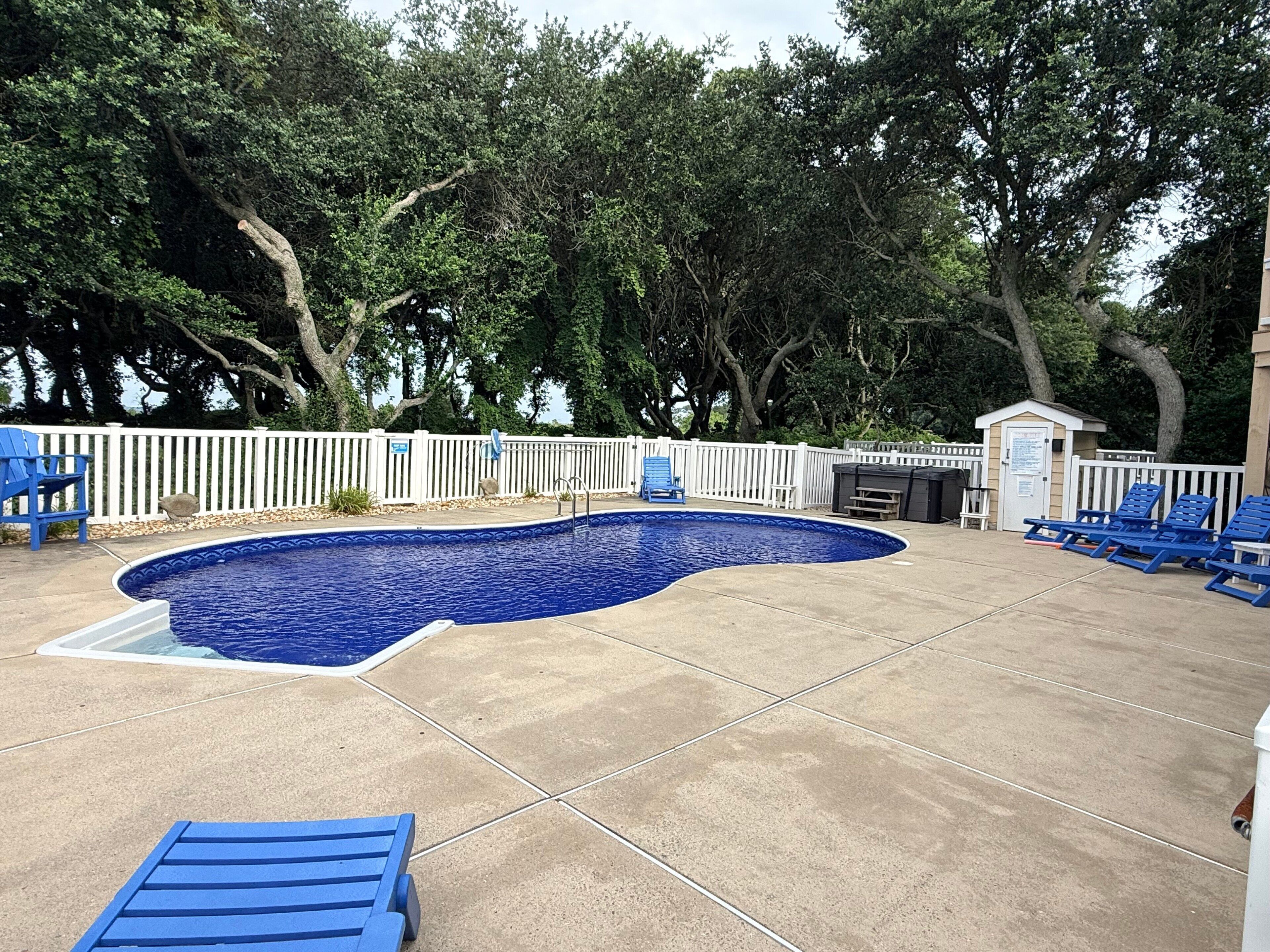 A swimming pool with a white fence, blue lounge chairs, and trees in the background.