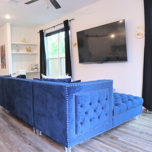 Un salon moderne avec un canapé d’angle bleu, un téléviseur à écran plat fixé au mur et une fenêtre ornée de rideaux noirs.