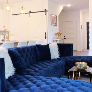 A modern living room with a blue sectional sofa, a small side table, and a dining area in the background. - New Gated Home Near the Texas Medical Center: Workspace + Yard +Fast Wifi (Houston)