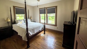 A four-poster bed with a white bedspread, a nightstand with a lamp, and a dresser with a television. - Build memories in WNC! (Brevard)