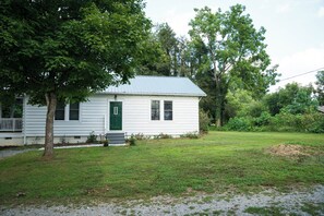 A single-story white house with a green door and windows, surrounded by a grassy yard and trees. - Build memories in WNC! (Brevard)