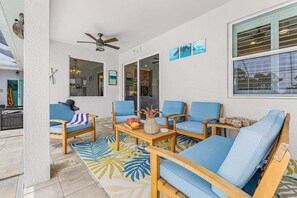 Eine sonnendurchflutete Terrasse mit blauen Polsterseats, einem Holztisch mit Obst und einem Deckenventilator.