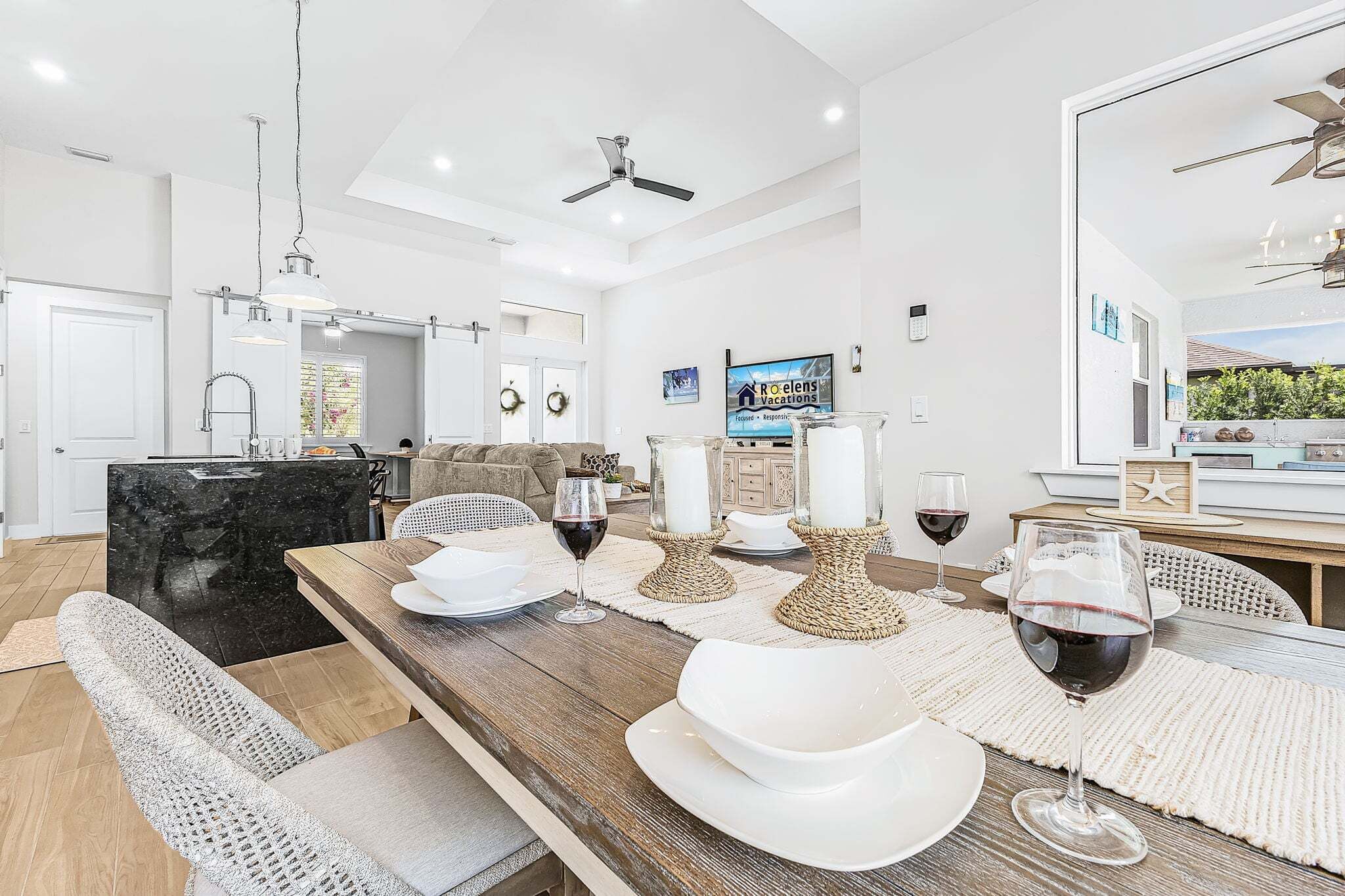 A modern dining area with a long wooden table set for a meal, a kitchen with a black marble countertop, and a living room with a sofa and a television.