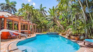 Une piscine entourée d’une terrasse en pierre, de chaises longues et d’une végétation tropicale.