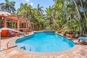 A swimming pool with a stone deck, lounge chairs, and tropical vegetation. - Tropical Retreat with Heated Pool, Spa & Tiki Bar - Coastal Paradise Getaway - Roelens (Cape Coral)