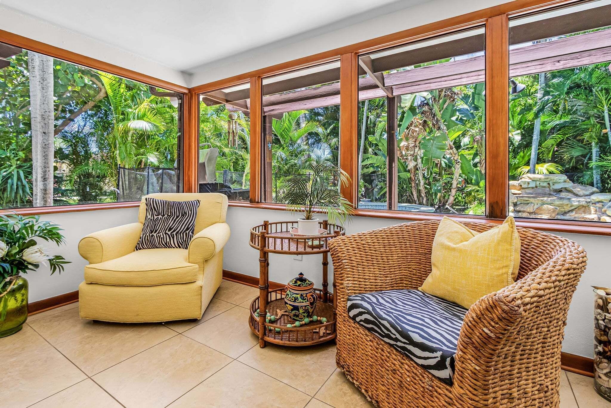 A room with a yellow armchair, a wicker sofa, and a small table with a decorative pot. Large windows offer a view of lush greenery outside.