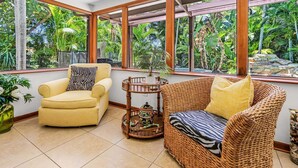 Une pièce avec un fauteuil jaune, un canapé en osier et une petite table sur laquelle est posé un pot décoratif. De grandes fenêtres offrent une vue sur la verdure luxuriante à l’extérieur.