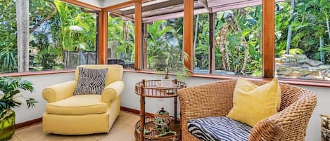 A room with a yellow armchair, a wicker sofa, and a small table with a decorative pot. Large windows offer a view of lush greenery outside.