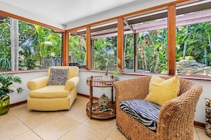 A room with a yellow armchair, a wicker sofa, and a small table with a decorative pot. Large windows offer a view of lush greenery outside. - Tropical Retreat with Heated Pool, Spa & Tiki Bar - Coastal Paradise Getaway - Roelens (Cape Coral)