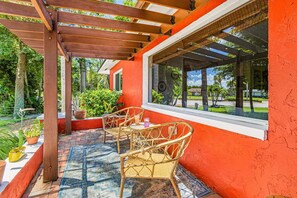 A covered outdoor seating area with wicker chairs and a table, a red wall, and a window overlooking a garden. - Tropical Retreat with Heated Pool, Spa & Tiki Bar - Coastal Paradise Getaway - Roelens (Cape Coral)