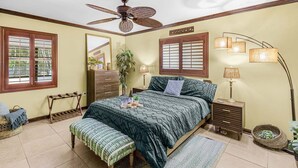 Une chambre à coucher comprenant un lit, des tables de chevet, un ventilateur de plafond, un miroir et une fenêtre avec des stores.