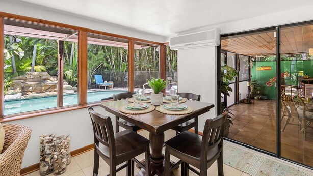 Un coin repas avec une table dressée pour quatre personnes, de grandes fenêtres et une vue sur une piscine et des plantes tropicales.