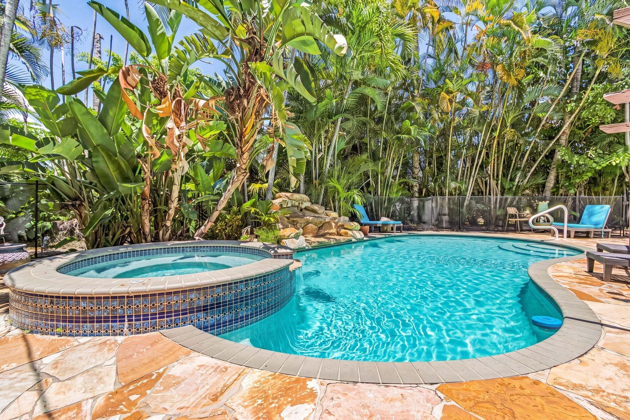 Piscina com hidromassagem, cercada por plantas tropicais e um deck de pedra.