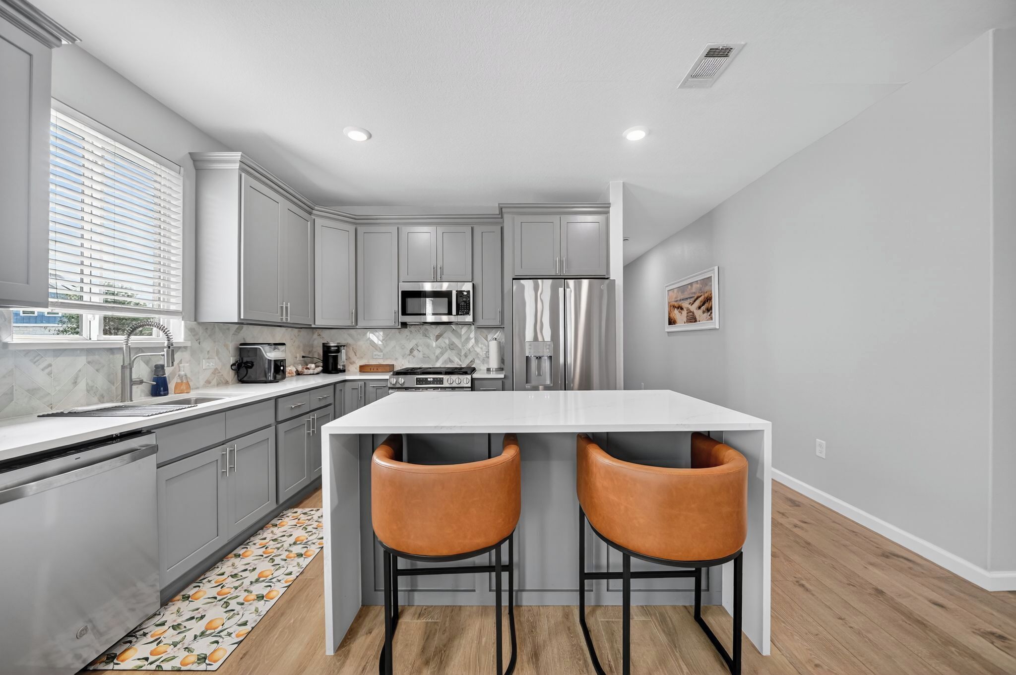Une cuisine moderne avec des meubles gris, un plan de travail blanc, un réfrigérateur en acier inoxydable et deux chaises orange.