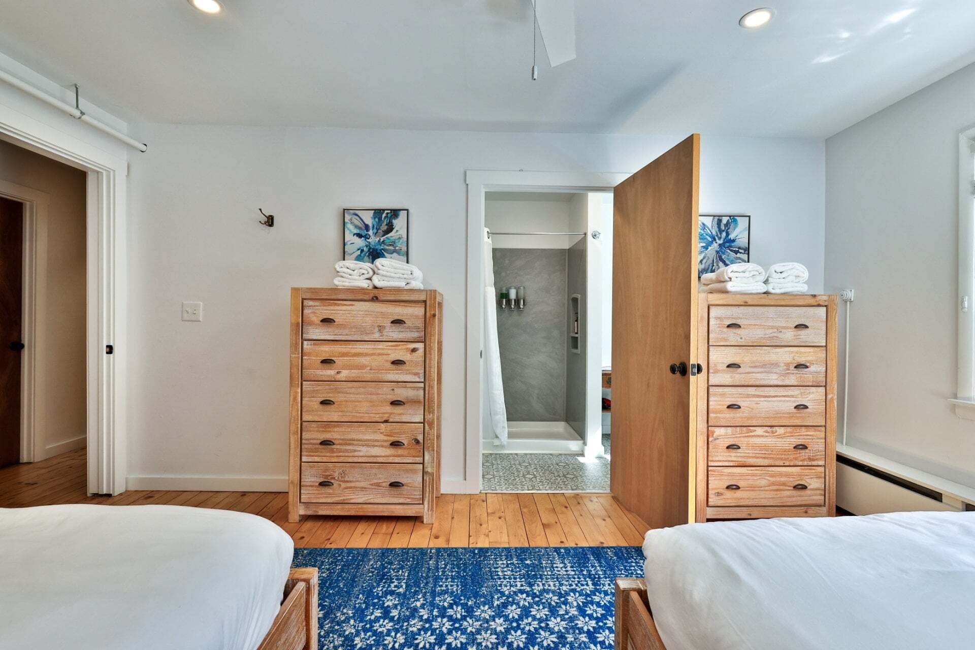 A bedroom with two beds, wooden dressers, a shower, and a blue patterned rug.