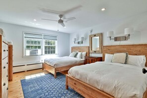 Une chambre avec deux lits, un ventilateur de plafond, une fenêtre avec des stores, une commode en bois et un tapis bleu à motifs.