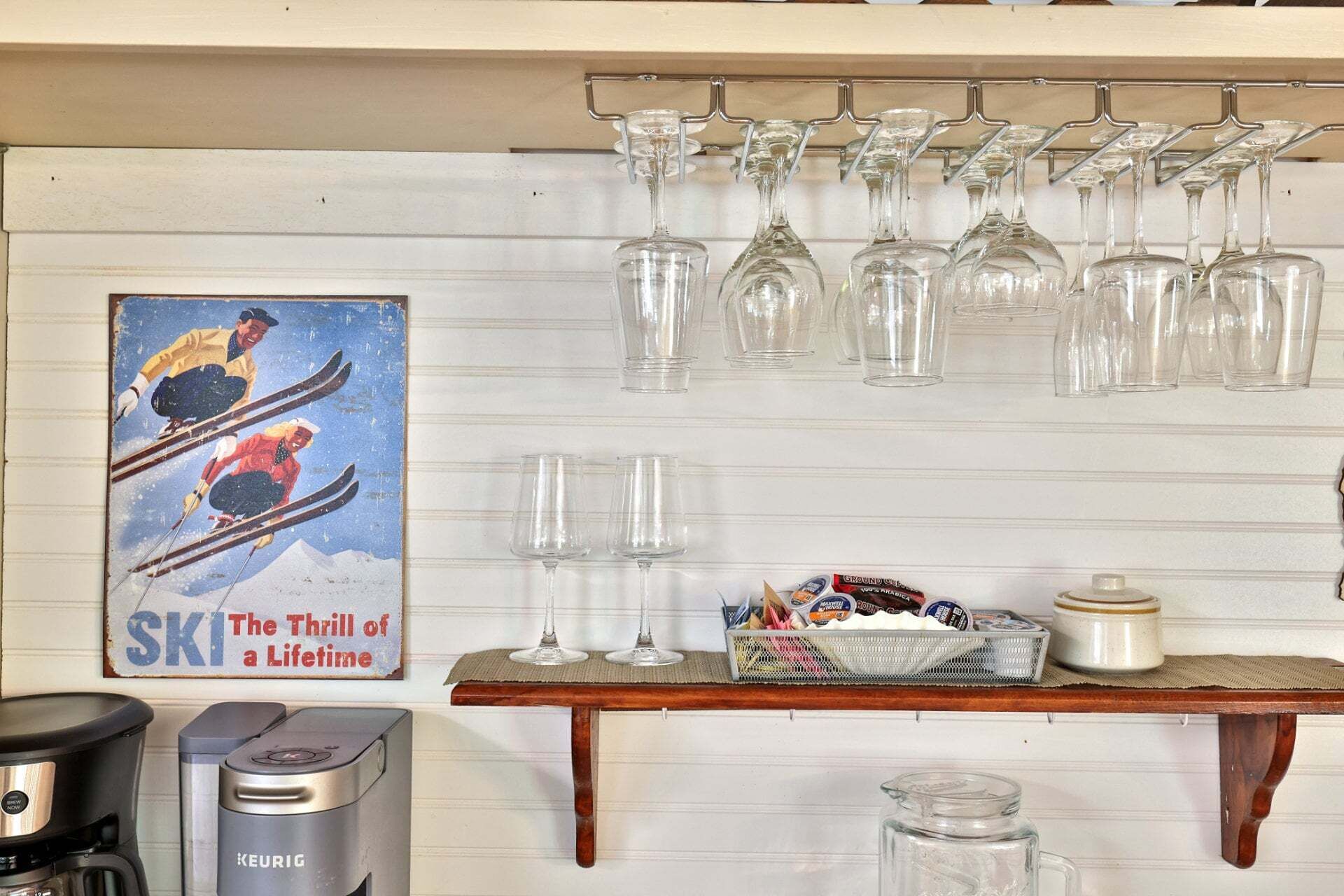 A kitchen with a coffee maker, a kettle, a shelf with wine glasses, a poster, and a basket with snacks.