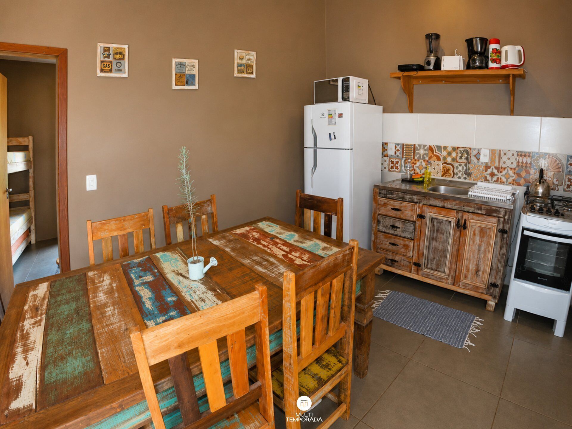 Una cocina con una mesa de madera, sillas, un refrigerador, una estufa y un estante con utensilios de cocina.
