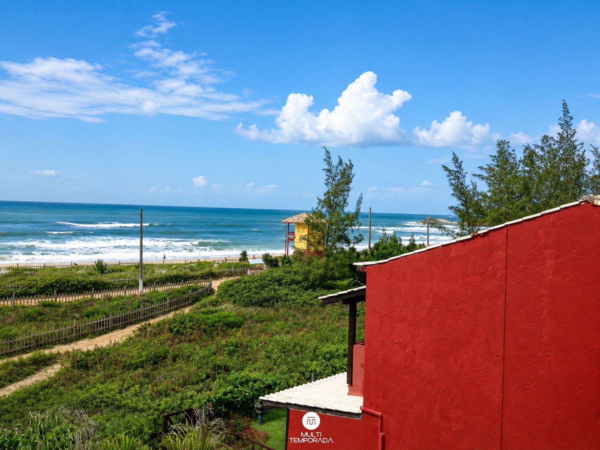 Edificio rojo con un letrero que dice 'MULTI TEMPORADA' frente a una playa con olas y cielo despejado.