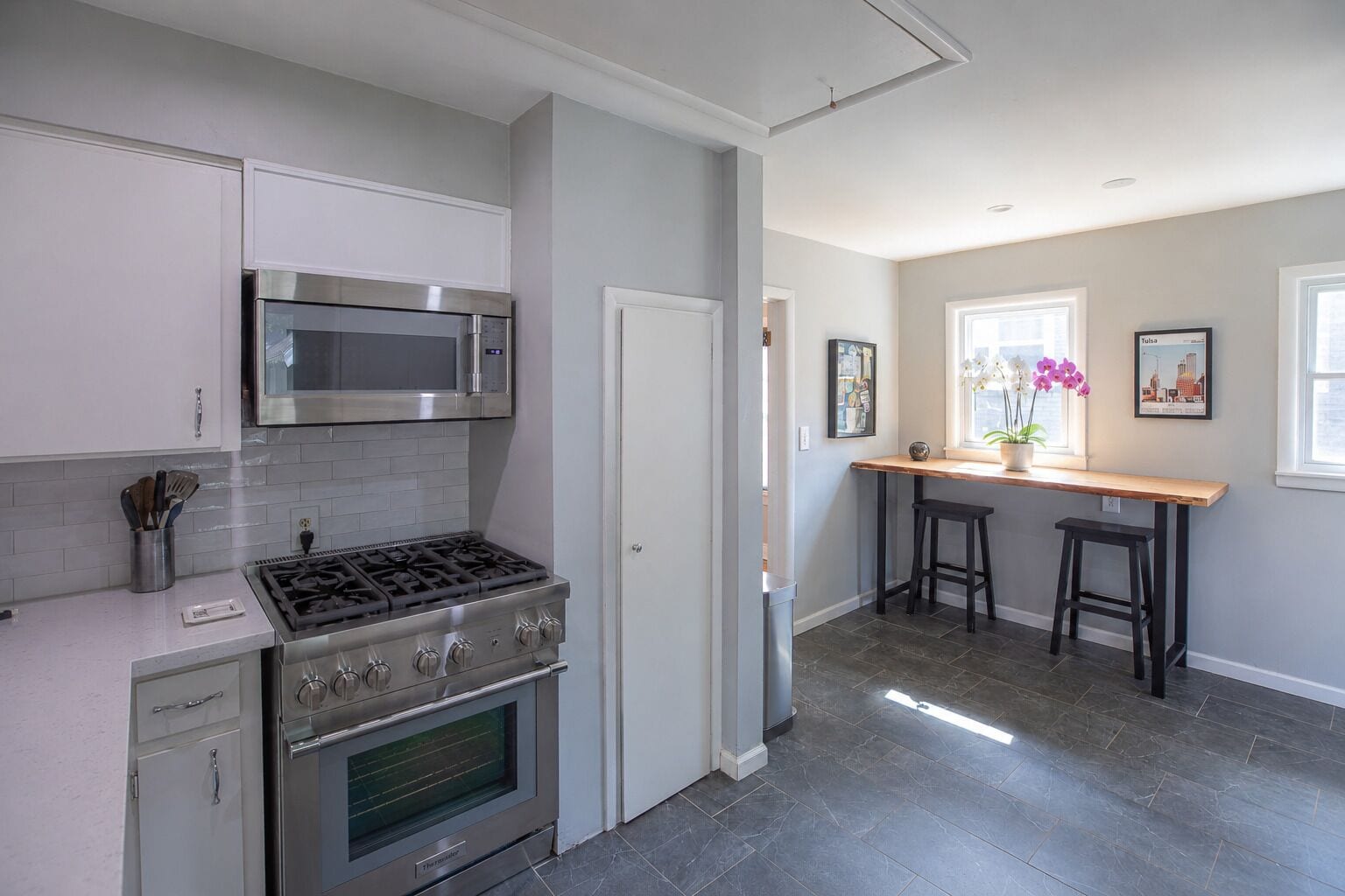 A modern kitchen with stainless steel appliances, a dining area with a wooden table and bar stools, and a view of the city through large windows.
